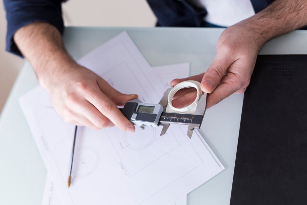 A person is using a digital caliper to measure a circular object, with technical drawings and a pencil visible on the desk.